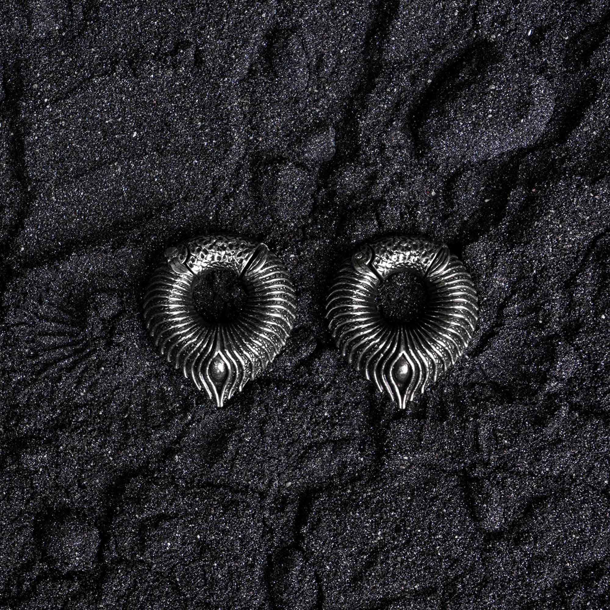 On a textured black surface lie two intricately designed, metallic, ring-shaped objects. Resembling curled, ornate serpents, these MARVDASHT Medium Cyborg Clicker Ear Weights from Kaya Movement feature detailed patterns with pointed ends touching the ground. Made of 925 silver and measuring 3/8" gauge, the dark and granular backdrop enhances their dystopian body jewelry aesthetic.